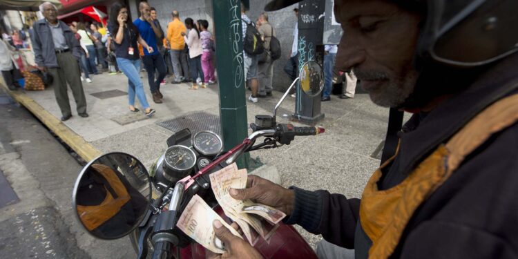 A man counts 100 Bolivar notes in the street whilst people queue outside a bank in Caracas in an attempt to deposit money, on December 15, 2016.
According to the UN's Economic Commission for Latin America and the Caribbean (ECLAC) forecast, Venezuela's economy will shrink 9.7 percent this year. / AFP PHOTO / FEDERICO PARRA