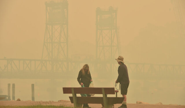 Tourists walk through dense smoke from bushfires in front of the Batemans Bay bridge as cars line up to leave the town in New South Wales to head north on January 2, 2020. - A major operation to move people stranded in fire-ravaged seaside towns was under way in Australia on January 2 after deadly bushfires ripped through popular tourist spots and rural areas leaving at least eight people dead. (Photo by PETER PARKS / AFP)