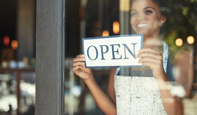 Cropped shot of a young woman hanging up an open sign on the window of her cafe