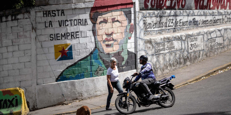 AME4413. CARACAS (VENEZUELA), 02/02/2024.- Personas pasan frente a un mural del ex presidente fallecido Hugo Chávez , el 26 de enero de 2024, en Caracas (Venezuela). Lo que tiene el chavismo en el poder lo tienen los venezolanos Gabriel Gutiérrez y Melannie Bonfissuto en edad: 25 años, por lo que solo han visto en el Gobierno de su país a un único sistema político que, pese a sus transformaciones en la última década, mantiene vivas la imagen y la palabra del fallecido Hugo Chávez. EFE/ Rayner Peña R.