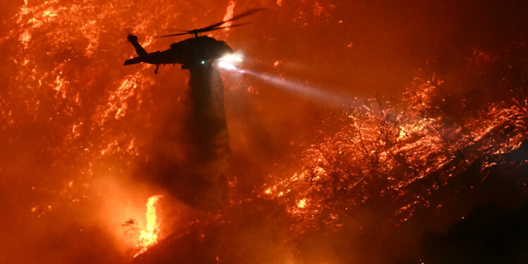 A fire fighting helicopter drops water as the Palisades fire grows near the Mandeville Canyon neighborhood and Encino, California, on January 11, 2025. The Palisades Fire, the largest of the Los Angeles fires, spread toward previously untouched neighborhoods January 11, forcing new evacuations and dimming hopes that the disaster was coming under control. Across the city, at least 11 people have died as multiple fires have ripped through residential areas since January 7, razing thousands of homes in destruction that US President Joe Biden likened to a "war scene." (Photo by Patrick T. Fallon / AFP)